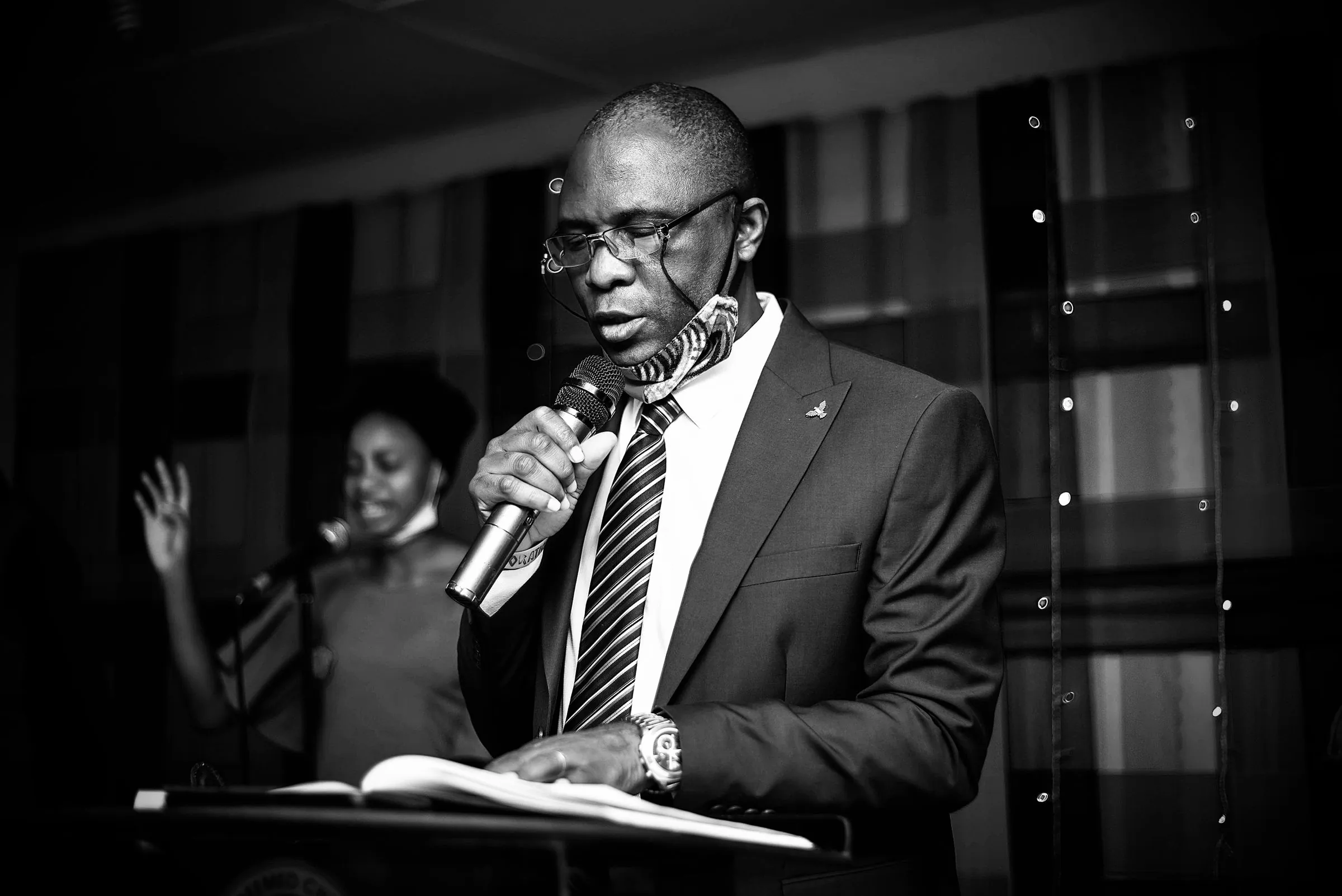 Black and white photo of a businessman in formal wear speaking into a microphone while reading a book.